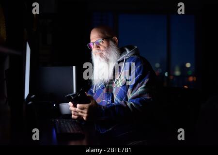 Mature bald bearded hipster man thinking and using phone at home late at night in the dark Stock Photo