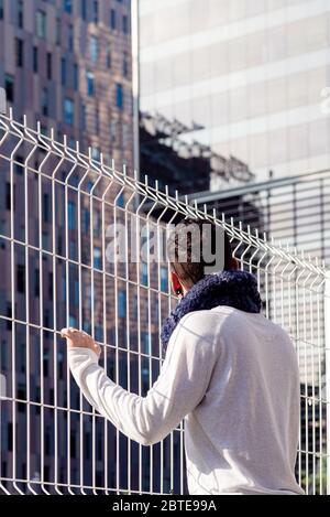Young male leaning on a metallic fence while looking away to the business buildings Stock Photo