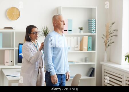 Side view portrait of female doctor listening to heart and breathing of senior patient, putting stethoscope on back during consultation in clinic, cop Stock Photo