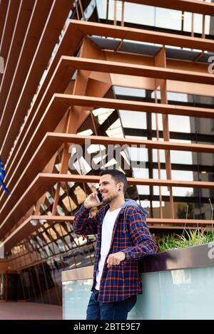 Young bearded male leaning on a wall wearing sunglasses while using a smartphone outside. Stock Photo