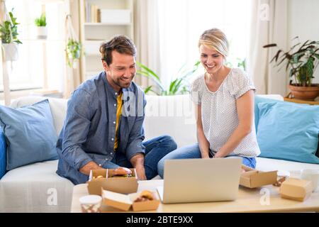 Happy couple with hamburgers sitting on sofa indoors at home, using laptop. Stock Photo