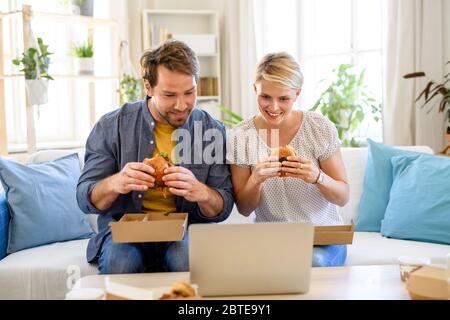 Happy couple sitting on sofa indoors at home, eating hamburgers. Stock Photo