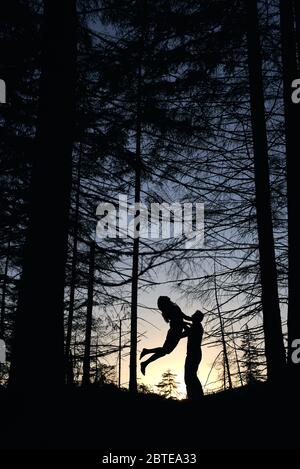 From below view of silhouettes of loving couple posing in evening forest. Wild nature with amazing views, summer sunset, man holding woman while jumping and feeling happy. Nature, love concept.