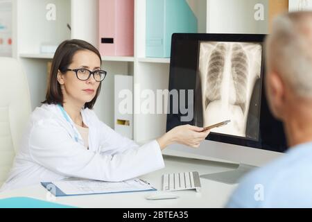 Portrait of serious female doctor pointing at x-ray image of lungs and chest while consulting senior patient in clinic Stock Photo