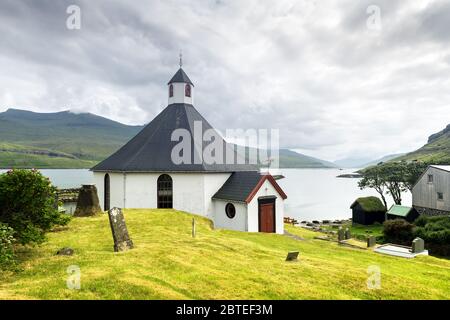 Summer view of traditional church in faroese village. Beauty landscape with foggy fjord and high mountains. Faroe Islands, Denmark. Stock Photo