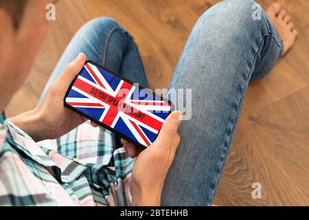 closeup of a young caucasian man, wearing casual clothes, sitting on a wooden floor, having his smartphone in his hands with the text do you speak Eng Stock Photo