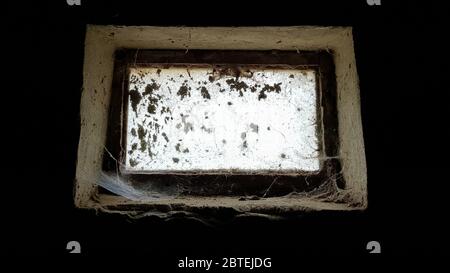 a small rectangular dirty and dusty web window in a large basement. White light penetrates the dark basement Stock Photo