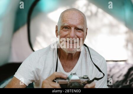 Closeup of a senior man sitting outside his camping tent with a digital camera. Retired man photographing while camping in nature. Stock Photo