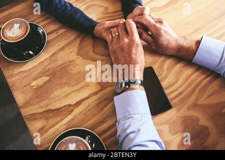 Close up of mature man and woman holding hands on a table with cups of coffee by the side. Top view of a senior couple sitting at a cafe.