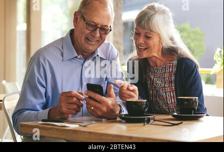 Young senior couple sitting at table in cafe and looking at mobile phone and smiling. Retired man and woman in coffee shop using smart phone. Stock Photo