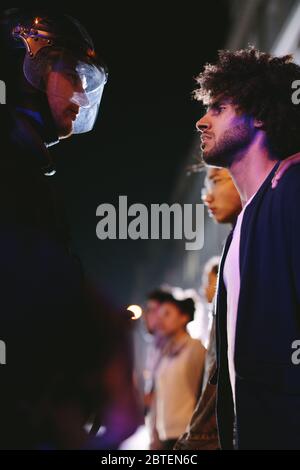 Male activist protesting against police violence. Young man looking into eyes of a policeman while standing with group of protestors at night. Stock Photo