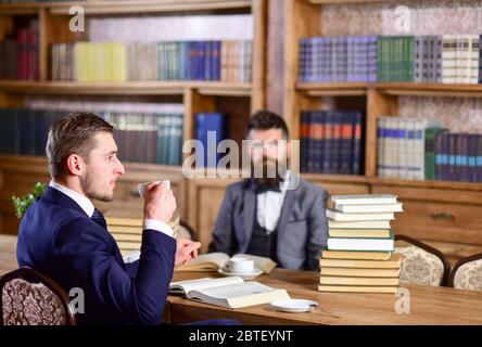 Men with busy faces drink tea. Men in suits, oldfashioned professors in library or vintage interior with bookshelves with antique books on background. Intellectuals and aristocrats concept. Stock Photo