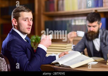 Men with busy faces reading books, studying and drink tea. Men in suits, aristocrats, professors in library or vintage interior with bookshelves on background. Intellectuals and aristocrats concept. Stock Photo