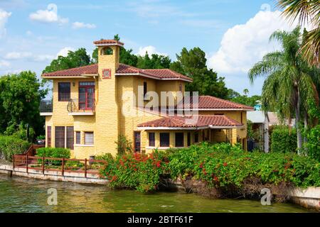 Yellow Italian style house with two stories and a chimney by the water edge with lush trees and flowering bushes on a beautiful sunny morning in the t Stock Photo