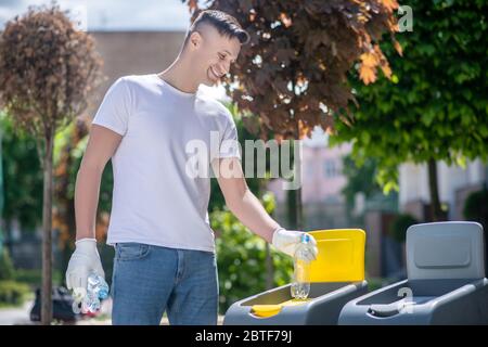 Dark-haired male in protective gloves throwing plastic bottle into waste container Stock Photo