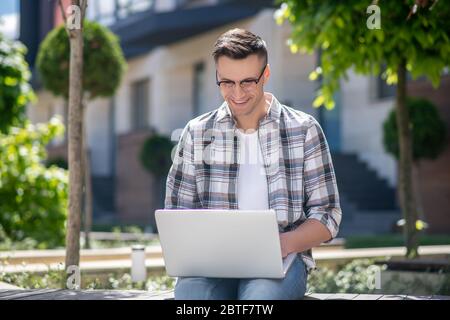 Dark-haired male wearing glasses, sitting in the yard and working on his laptop Stock Photo