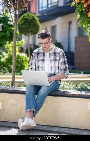 Dark-haired male wearing glasses, sitting on bench and working on his laptop Stock Photo