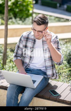 Dark-haired male sitting in the yard and working on his laptop, touching his glasses with one hand Stock Photo