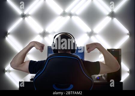Back view of young gamer guy in headphones screaming and rejoicing unleash power while playing video games on computer young man shows his biceps Stock Photo