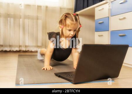 4 years old girl stands in a plank pose in front of a laptop at home, sunlight, backlight. online gymnastics classes Stock Photo