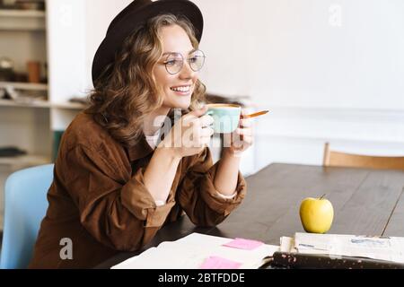Image of joyful charming woman in eyeglasses drinking coffee while studying with exercise books at home Stock Photo