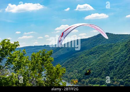 A man flying a white paraglide over the forest and the river on a beautiful sunny day. Stock Photo