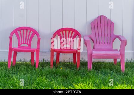 Three miniature children's colourful plastic chairs on the lawn against a white wooden fence Stock Photo