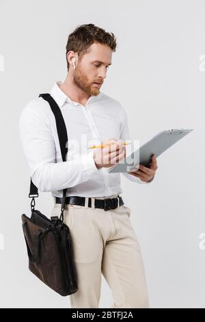 Image of focused man in wireless earphones holding clipboard and writing isolated over white background Stock Photo