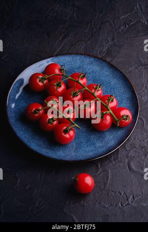 Cherry tomato branch, fresh ripe organic vegetables in blue plate on dark black textured background, angle view Stock Photo