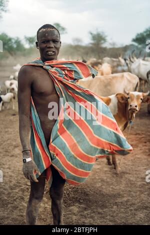MUNDARI TRIBE, SOUTH SUDAN - MARCH 11, 2020: Young man shepherd in colorful waving clothes looking at camera with distrust while standing against herd Stock Photo