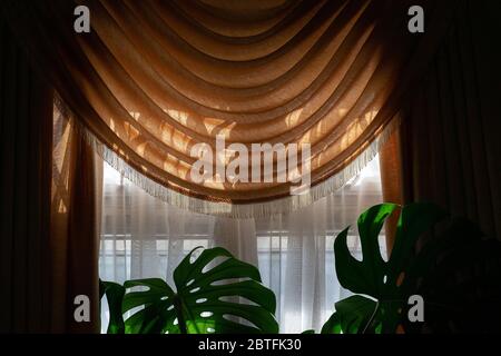 Part of a room window with translucent thin white and a luxurious draped orange curtain at the top and sides against the evening sunlight Stock Photo