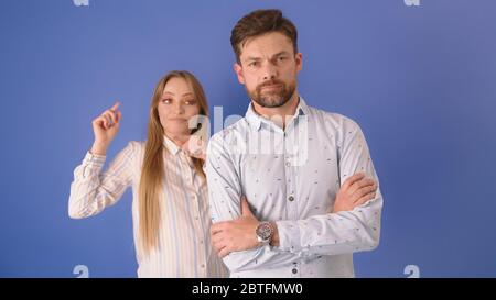 Man and the woman isolated on the blue background. Woman using sign language behind mans back. Stock Photo