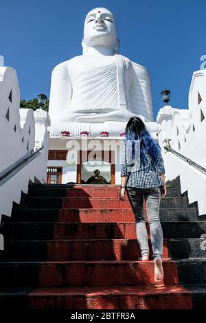 Blue haired girl going barefoot up the stairs to the white Buddha monument Bahirawakanda in Kandy, Sri Lanka. Visiting a peaceful temple and exploring Stock Photo