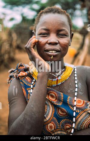 TOPOSA TRIBE, SOUTH SUDAN - MARCH 12, 2020: Young woman with traditional scars of Toposa Tribe touching face and looking at camera on blurred Stock Photo