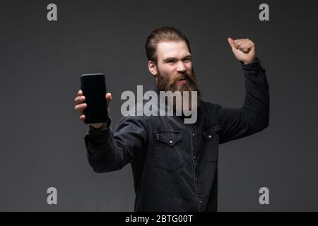 Portrait of man holding phone scream with fist up on over gray background Stock Photo