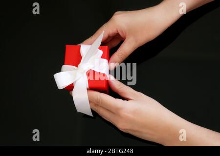 Red gift box in female hands on dark glass background. Woman holds present tied by white ribbon, concept of birthday, surprise, anniversary Stock Photo