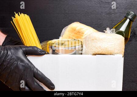 Close up mans hands in black rubber gloves holding a white box with pasta, cereals and canned food on a black background. Safety food delivery service Stock Photo