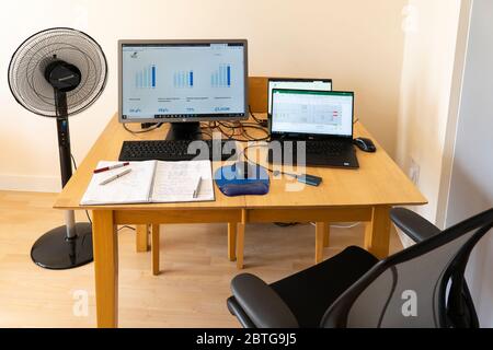 A table with laptops and a monitor with a mouse and mouse mat, notepad and pens, a fan, and screens showing graphs. Theme - working from home. UK Stock Photo