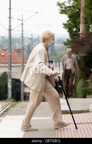 Belgrade, Serbia - May 21, 2020: Senior woman wearing face surgical mask walking with a cane on zebra crossing Stock Photo