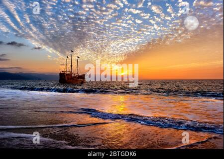 An Old Wooden Pirate Ship Sits on the Ocean at Sunset as the Full Moon rises in the Sky Stock Photo