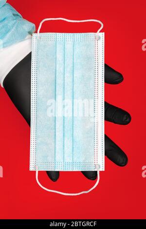 Paramedic holding blue surgical face mask in hand black protective glove. Medical, epidemic and hygiene concept on bright red background Stock Photo