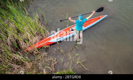 senior male paddler is stretching before workout on his racing stand up paddleboard, aerial view of a lake shore Stock Photo