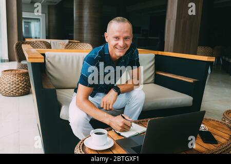 A young smiling guy is reading mail on a laptop, Look into the camera, sitting in a spacious cafe with a cup of coffee. The student makes an entry in Stock Photo