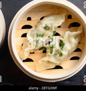 Gyozas potstickers chinese dumplings in wooden steamer with soy sauce fresh herbs and chopsticks on old wood tabletop view overhead close up nobody. Stock Photo