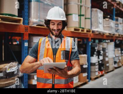 Successful entrepreneur in warehouse checking inventory list using digital tablet while wearing helmet and safety vest for protection Stock Photo