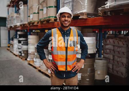 Portrait of happy manager standing with his hands on waist wearing white helmet and safety vest in warehouse with shelves of inventory in the Stock Photo