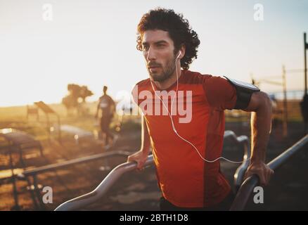 Portrait of an athletic young man listening to music on earphones doing exercise on parallel bars in the park - young man doing dips outdoors Stock Photo