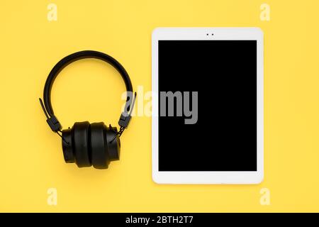 headphones, white tablet with black screen isolated on yellow background Top view Flat lay Listening to music online, audio books, training course Stock Photo