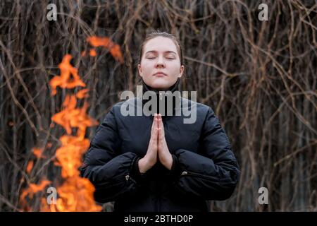 A young girl in a black jacket with hands clasped in prayer and closing her eyes is standing by the fire. Bright orange bonfire flames. Stock Photo