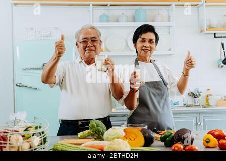 Happy Asian senior couple drinking milk at home. 70s Elderly married man and woman giving a thumbs up and holding a glass of milk at kitchen. Stock Photo